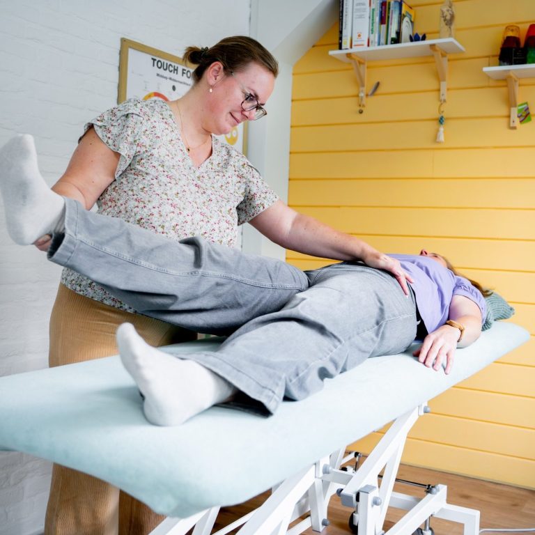 Een vrouw biedt een therapeutische behandeling aan een liggende patiënt in een lichte kamer. Kinesiologiebehandeling